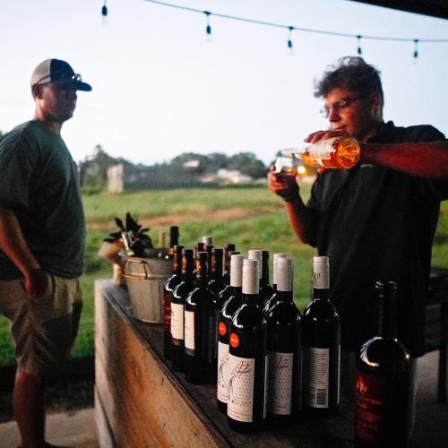 A person pours wine from a bottle while another stands nearby, with a scenic outdoor setting in the background.