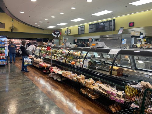 A grocery store deli section with various meats and cheeses, shoppers browsing, and a display of fresh bread.