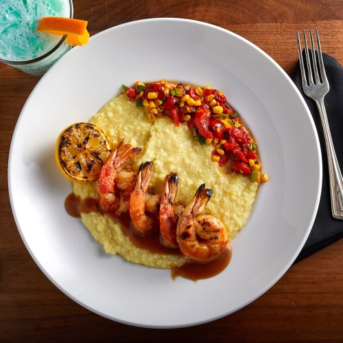 A plate of shrimp and grits topped with grilled shrimp, corn salsa, and a lemon slice, alongside a colorful drink.