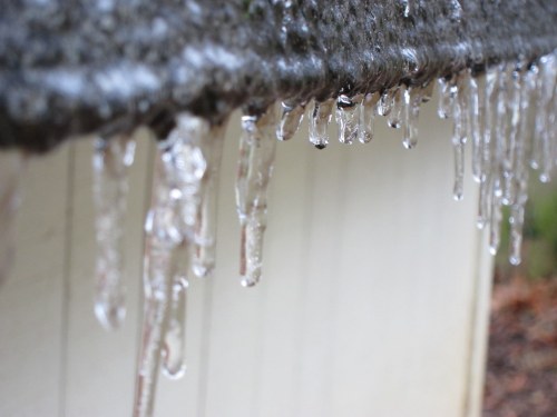 Icicles hang from a roof edge, glistening in the light against a blurred white background.
