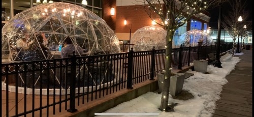 Outdoor dining igloos on a snowy patio, surrounded by string lights and a fence, with people inside enjoying the evening.