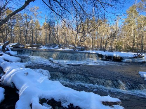 A serene winter scene of a river with a small waterfall, surrounded by snow-covered banks and bare trees.