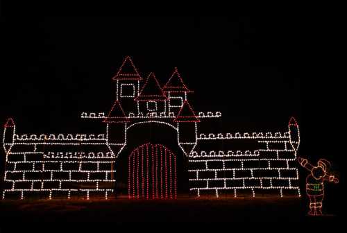 A brightly lit castle made of holiday lights, featuring towers and a red gate, with a festive figure beside it.