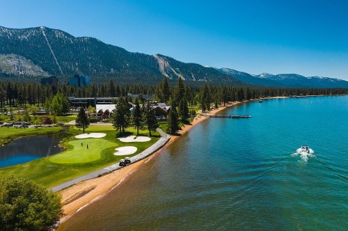 A scenic view of a golf course by a lake, surrounded by mountains and trees, with a boat on the water.