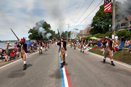 A parade with soldiers in historical uniforms marching along a road, while spectators watch from the sidelines.
