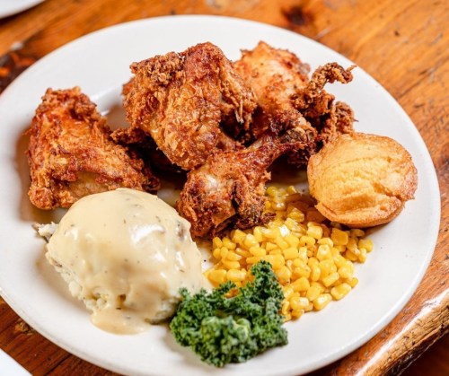 A plate of fried chicken, cornbread, mashed potatoes with gravy, corn, and a garnish of kale on a wooden table.