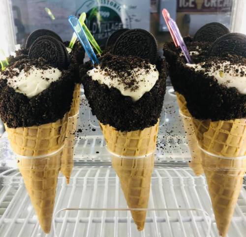 Four ice cream cones topped with whipped cream, crushed cookies, and colorful spoons, displayed in a freezer.