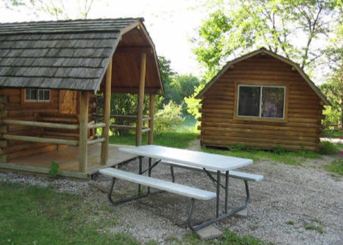 Two rustic wooden cabins with a picnic table in front, surrounded by greenery and trees.