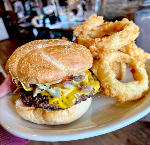 A hand holding a plate with a cheeseburger topped with lettuce and tomato, alongside crispy onion rings.
