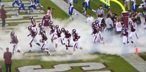 A football team runs onto the field through smoke, with fans and officials in the background.