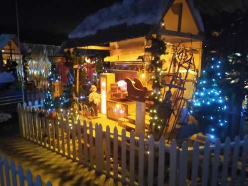 A cozy, illuminated holiday scene with a decorated house, trees, and a white picket fence, set at night.