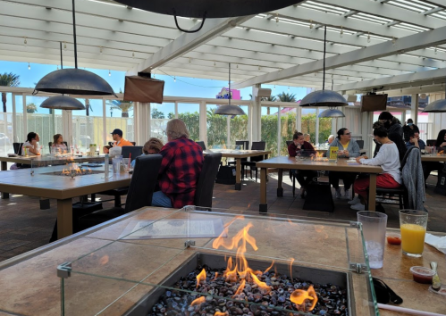 A lively restaurant with outdoor seating, featuring tables with fire pits and diners enjoying their meals.