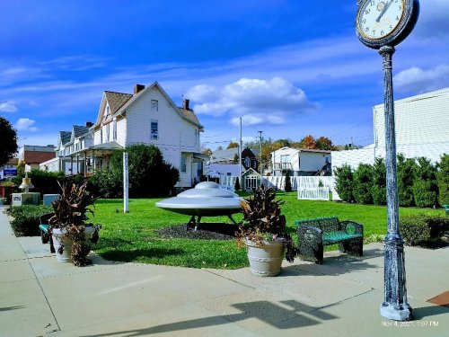 A park with a UFO sculpture, surrounded by green grass, potted plants, and a vintage clock in the foreground.