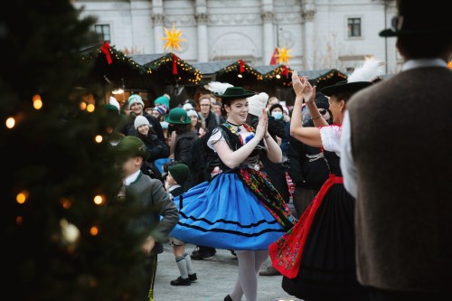 A festive scene with dancers in traditional costumes performing at a holiday market, surrounded by a cheerful crowd.