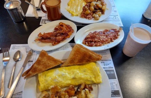 A breakfast spread featuring an omelet, bacon, toast, hash browns, and a drink on a table with utensils.
