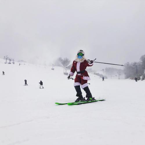 A child in a Santa costume skiing down a snowy slope, smiling and waving. Other skiers are visible in the background.