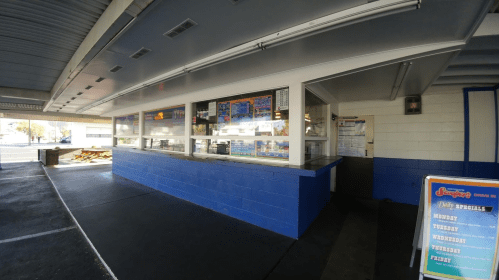 A drive-in food stand with a blue and white exterior, featuring a menu board and large windows for ordering.
