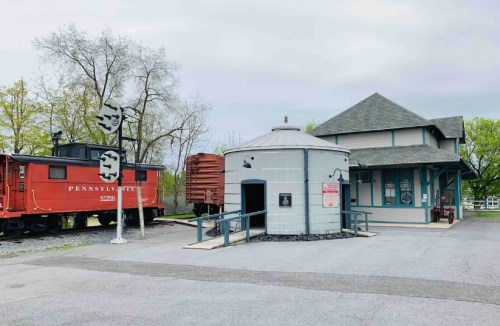 A red train car next to a round building and a historic train station, surrounded by trees and a cloudy sky.