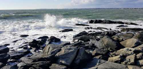 Waves crash against rocky shoreline under a cloudy sky, with distant land visible on the horizon.