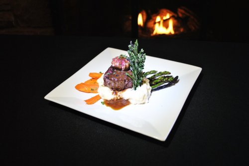A gourmet plate featuring steak, mashed potatoes, asparagus, and carrots, with a warm fireplace in the background.