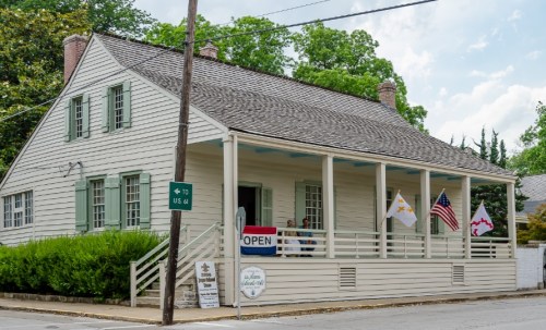 A historic house with a porch, flags, and an "Open" sign, surrounded by greenery on a sunny day.