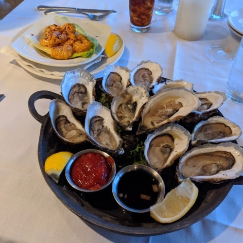 A platter of fresh oysters on ice with lemon wedges and dipping sauces, alongside a plate of shrimp in the background.