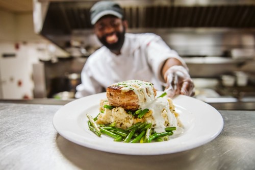 A chef presents a plate of chicken with creamy sauce, mashed potatoes, and green beans in a restaurant kitchen.