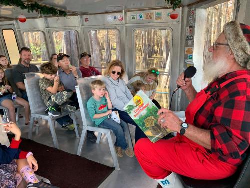 A man in a Santa outfit reads to a group of children and adults on a boat, surrounded by trees.