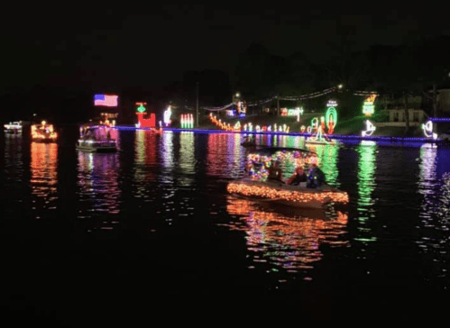 Colorful boats decorated with lights float on a dark lake, reflecting festive decorations along the shoreline.