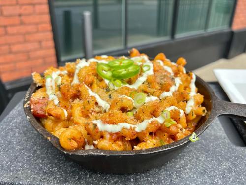 A cast iron skillet filled with pasta topped with green onions, sauce, and crispy bits, set against a brick background.