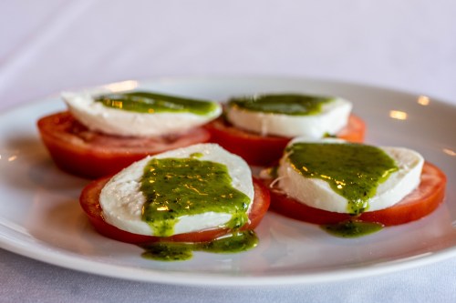 Sliced tomatoes topped with fresh mozzarella and drizzled with green pesto on a white plate.