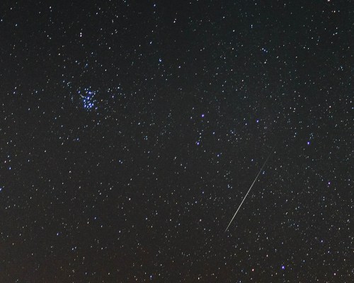A starry night sky filled with numerous stars, featuring a bright meteor streaking across the scene.
