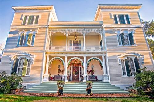 A large, elegant two-story house with a porch, decorative arches, and festive decorations. Lush greenery surrounds the entrance.