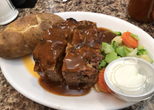 A plate featuring meatloaf with gravy, a baked potato, and a side of mixed vegetables and sour cream.