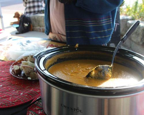 A close-up of a slow cooker filled with orange soup, with a ladle resting inside, on a table with a decorative cloth.