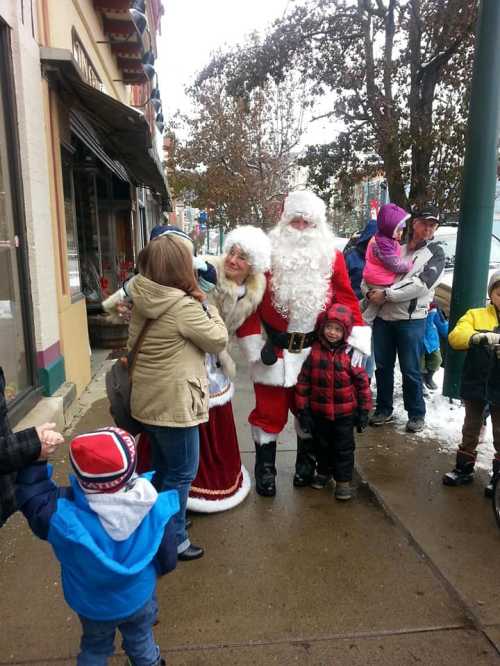 A festive scene with Santa and Mrs. Claus greeting children on a snowy street, surrounded by families and holiday cheer.