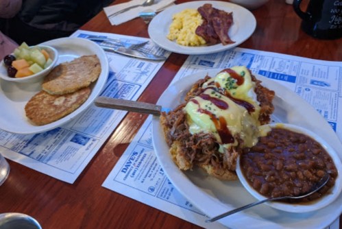 A plate with pulled pork topped with hollandaise sauce, baked beans, and a side of scrambled eggs and fruit.