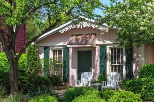 Charming pink cottage with green shutters, surrounded by lush greenery and two white chairs on the porch.