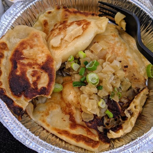 A foil container with golden-brown dumplings topped with chopped onions and green onions. A black fork is beside it.