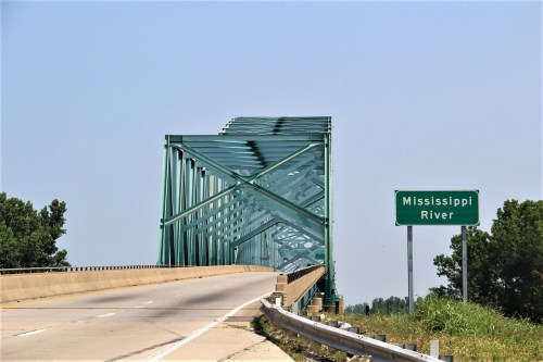 A green metal bridge spans the Mississippi River, with a road sign indicating the river's location nearby.