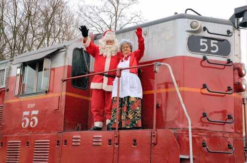 Santa Claus and Mrs. Claus wave from the platform of a red train engine, surrounded by trees.