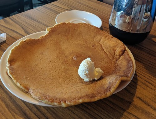 A large, golden-brown pancake topped with a dollop of butter, served on a white plate with a coffee pot in the background.