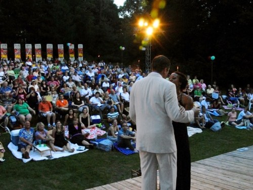 A couple performs on stage in front of a large audience seated on the grass, enjoying an outdoor event at dusk.
