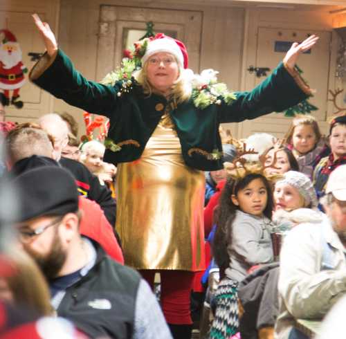 A festive crowd with a woman in a Christmas outfit, arms raised, surrounded by children and holiday decorations.