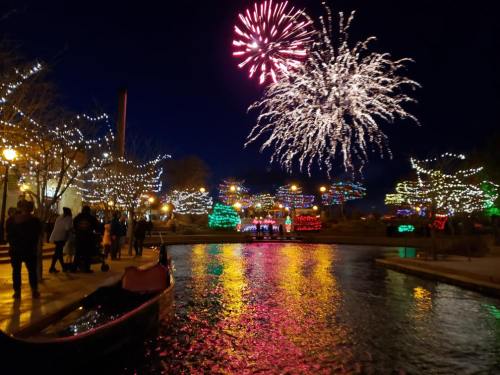 Colorful fireworks illuminate the night sky over a festive waterfront, with trees adorned in lights and people enjoying the scene.