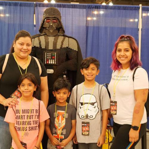 A group of four children and two adults pose with a large LEGO Darth Vader at a convention.