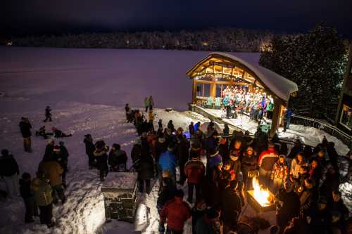 A crowd gathers around a snowy outdoor stage, enjoying a performance under a night sky with trees in the background.