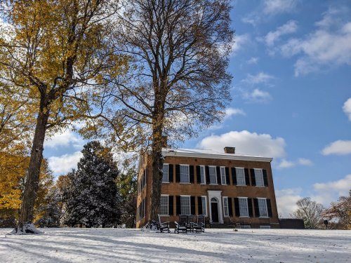 A historic house surrounded by trees, with a light dusting of snow and autumn leaves under a partly cloudy sky.