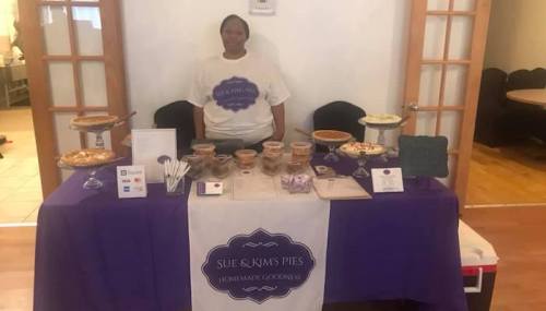 A woman stands behind a table displaying various homemade pies and desserts, with a purple tablecloth and signage.