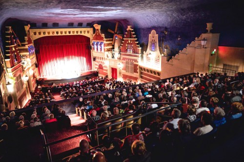 A packed theater with a red curtain, stage lights, and an audience seated in rows, enjoying a performance.